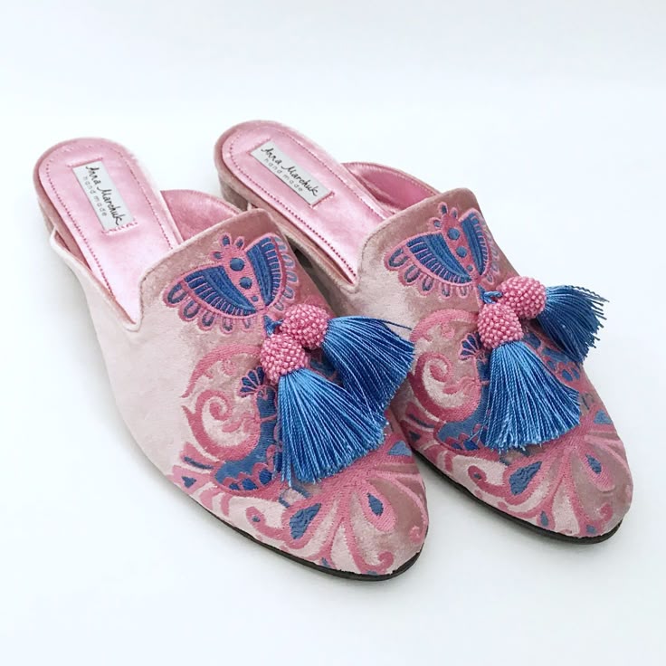 two pink velvet mules with blue and pink embroidery and blue tassels sit on a white background. the mules are openbacked and have a pink lining. the embroidery is intricate and detailed. the tassels are attached to the front of the mules.