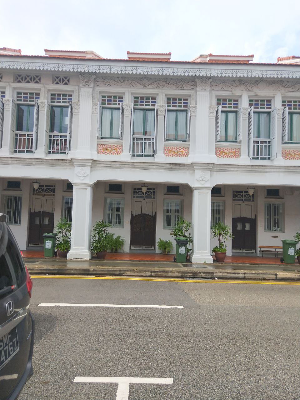 a shot of a white shophouse building in singapore. there is a car parked in front of the building with a parking spot marked in front of it. the building has many windows with green shutters and a few potted plants on the sidewalk.