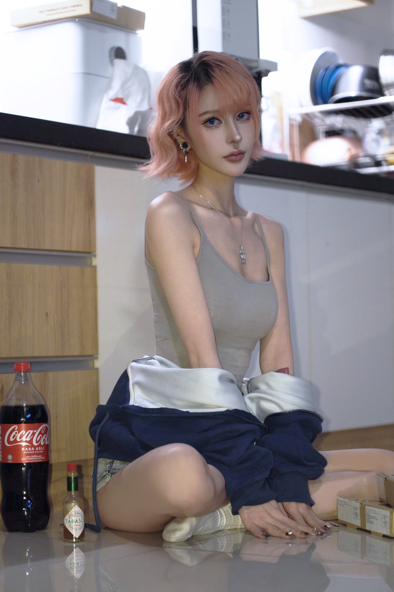 a young asian woman with short, pink hair sits on a kitchen floor. she is wearing a gray tank top, jeans shorts, a navy blue and gray hoodie, and white socks.  she has makeup on and is accessorized with earrings and a necklace. a bottle of cocacola and a bottle of hot sauce are on the floor next to her.  the kitchen cabinets and counter are visible in the background.