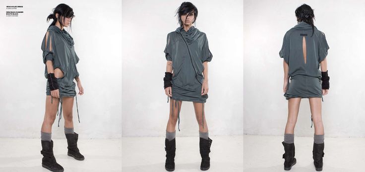 a woman stands in a studio, wearing a grey dress, black boots and black arm warmers. her hair is dark and she is posing with her hands on her hips.