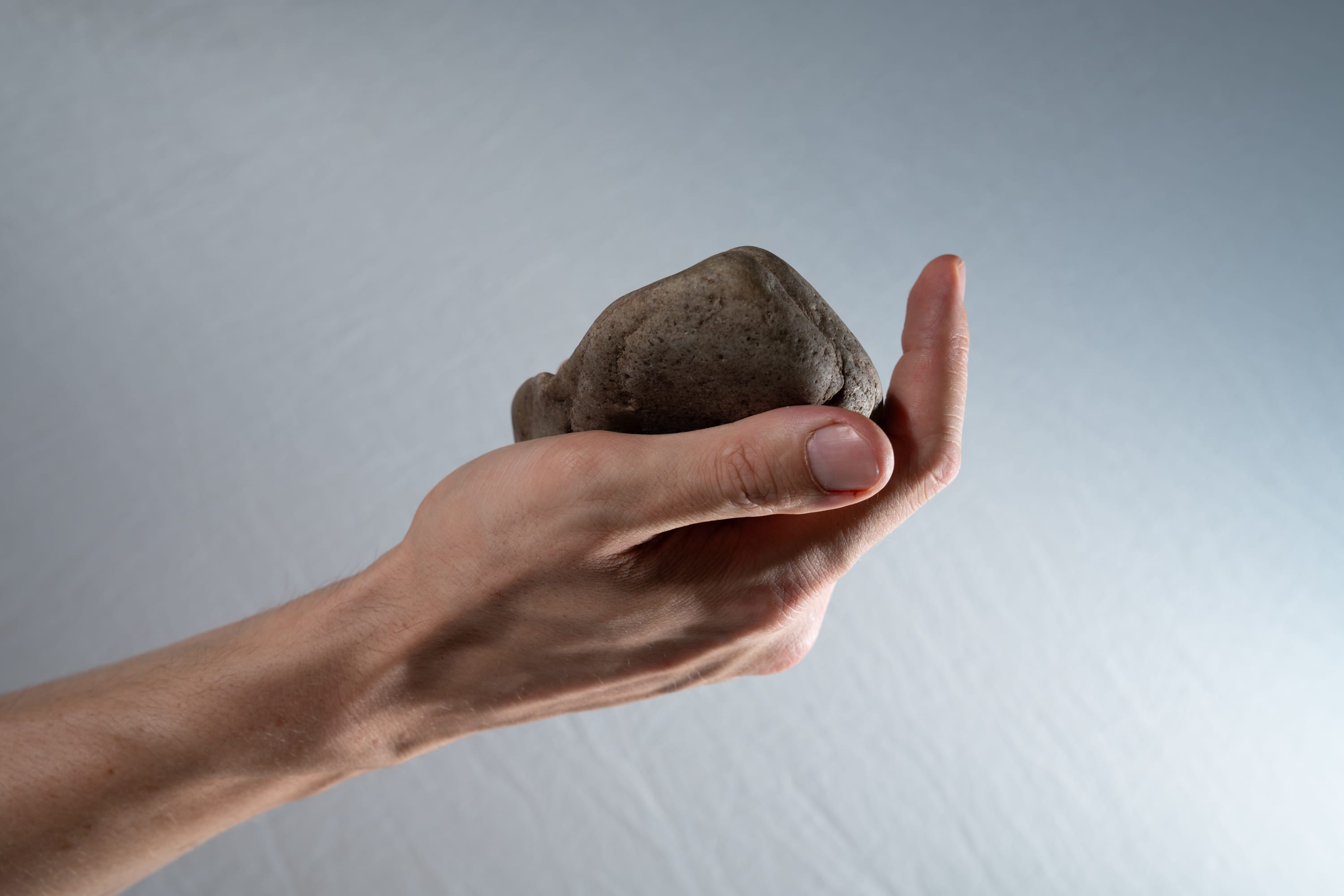 an adult hand holds a graybrown rock against a light gray backdrop. the rock appears rough and textured. the image is illuminated with studio lighting, creating a clear and focused view of the rock and hand.