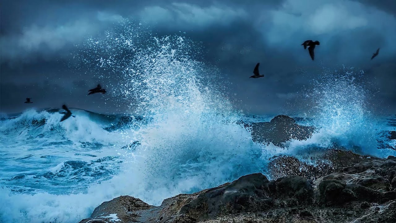 the image captures a dramatic scene of a stormy ocean. large, powerful waves crash against a rocky shoreline, sending plumes of white spray into the air. dark, heavy clouds fill the sky, casting a moody atmosphere over the scene. several birds are silhouetted against the churning water and the dark sky, adding to the sense of wildness and energy. the image is taken from an eyelevel perspective, placing the viewer directly in the midst of the storm's power.