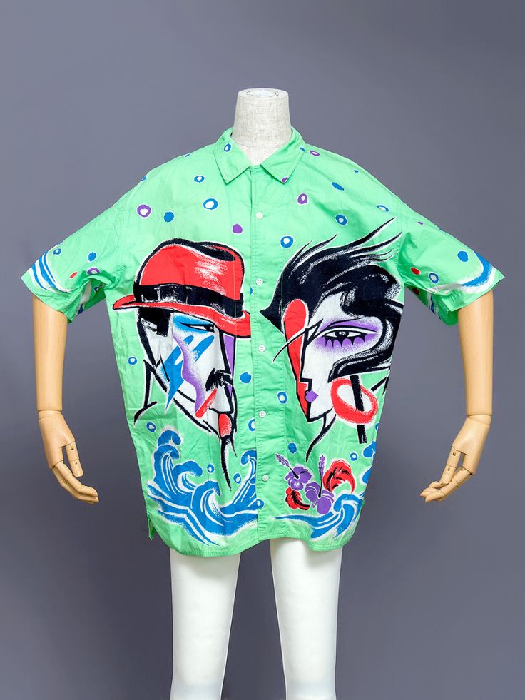 a green buttondown shirt with a vibrant and artistic pattern is displayed on a mannequin. the shirt features two abstract faces, one with a red hat and the other with stylized black hair, along with wavy blue lines at the bottom and colorful polka dots scattered throughout. the mannequin is white and partially visible against a gray background.