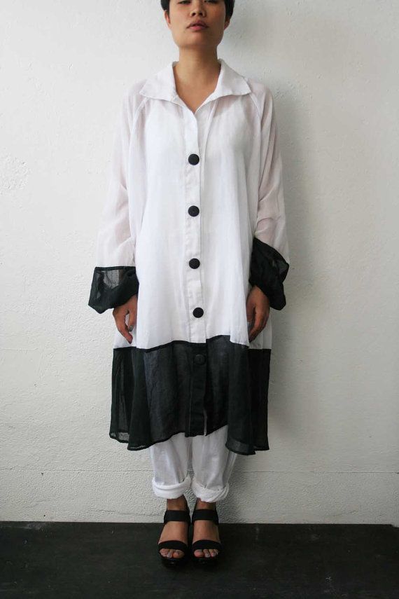 a full shot of a woman standing against a solid background, wearing a white, longsleeved shirt dress with black accents and black sandals. she also wears white pants. the dress has large black buttons and black detailing on the cuffs and hem. her hair is short and dark. she has a neutral expression.