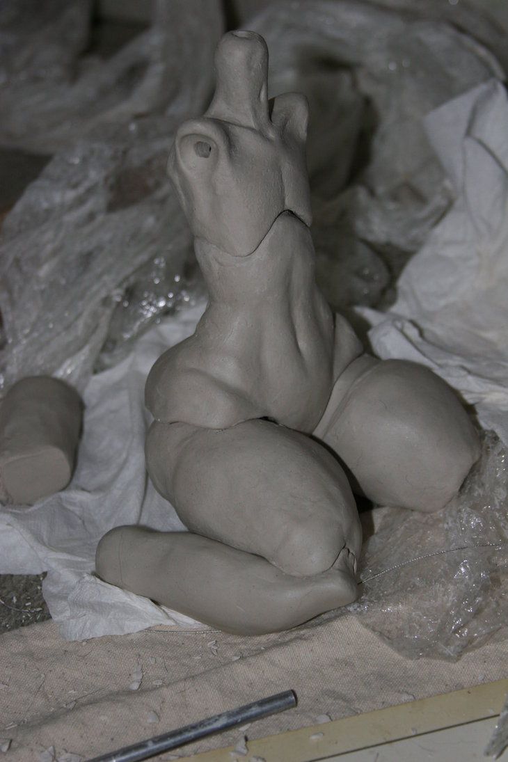 a closeup photo of a clay sculpture of a human figure, likely in the process of being made. the sculpture is sitting on a table covered in white fabric. the sculpture is in an unfinished state, with the clay still visible and slightly uneven. the photo is taken from a slightly high angle, giving a good view of the sculpture's form.