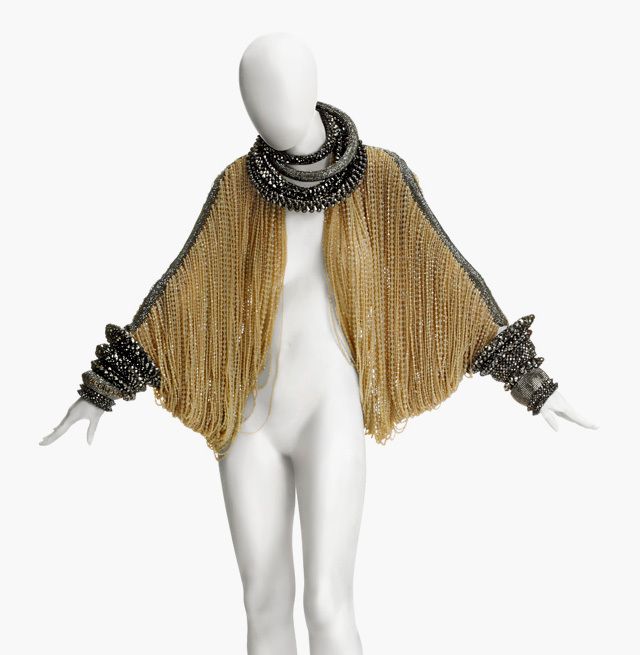 a mannequin is wearing a gold fringe jacket with black embellishments and a black necklace. the jacket has a batwing or capelike shape with dense gold fringe covering the body. the edges of the jacket, as well as the cuffs, are adorned with black embellishments, possibly beads or sequins. the necklace is a multilayered, chunky piece, also in black. the mannequin itself is a standard white, faceless model with its arms outstretched to better showcase the jacket. the background is a plain white, drawing full attention to the garment and accessories.