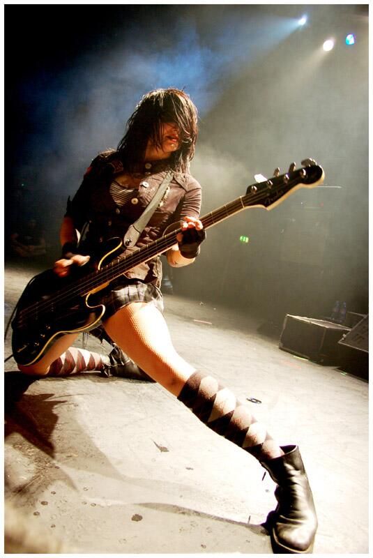a female rock star performs on stage, playing a bass guitar with energy and passion. the stage is dark and smoky with spotlights illuminating the musician. the woman is dressed in a black leather jacket, black miniskirt, and kneehigh patterned socks and boots.