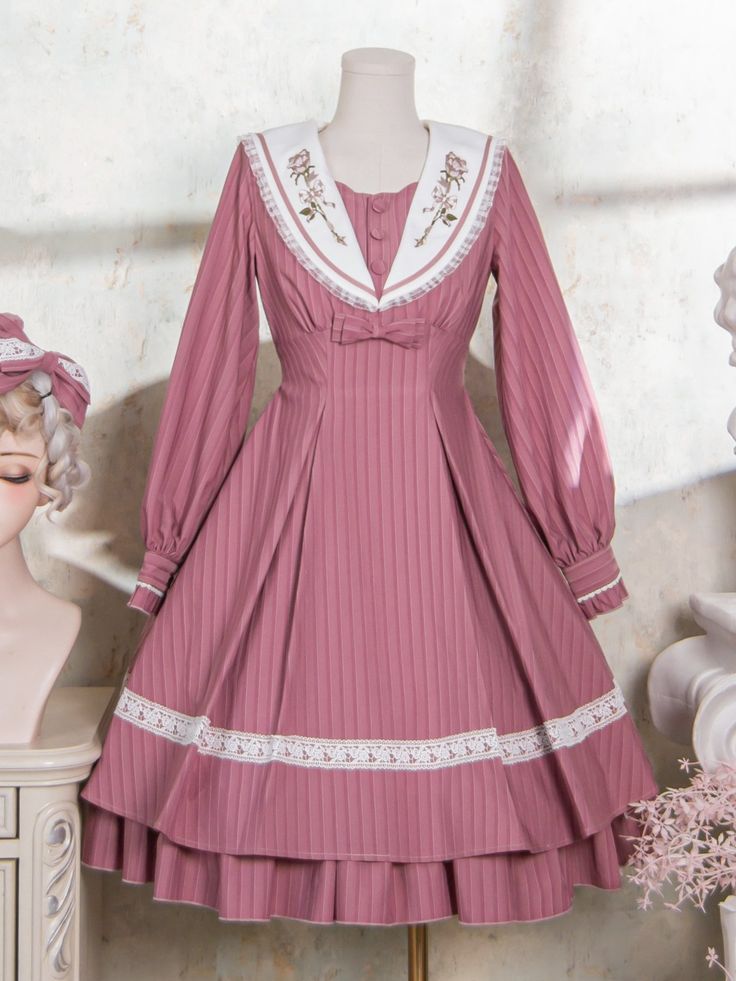 a fulllength shot of a pink dress with a sailor collar displayed on a mannequin. the dress is a vintageinspired style with delicate floral embroidery on the collar and lace trim. the mannequin is positioned against a white wall with a floral arrangement to the right. the lighting is soft and diffused, highlighting the intricate details of the dress.
