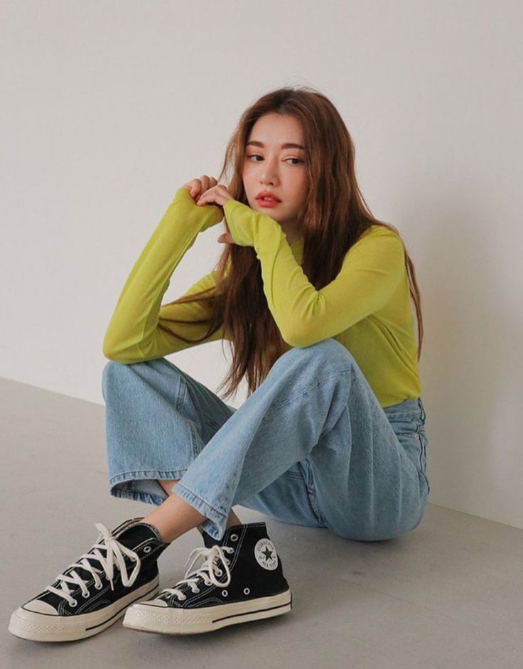 a young asian woman wearing a yellow long sleeve shirt and blue jeans is sitting on the floor with her legs crossed. she is wearing black high top converse shoes and her long brown hair is flowing down her back. she is looking to the left of the camera.