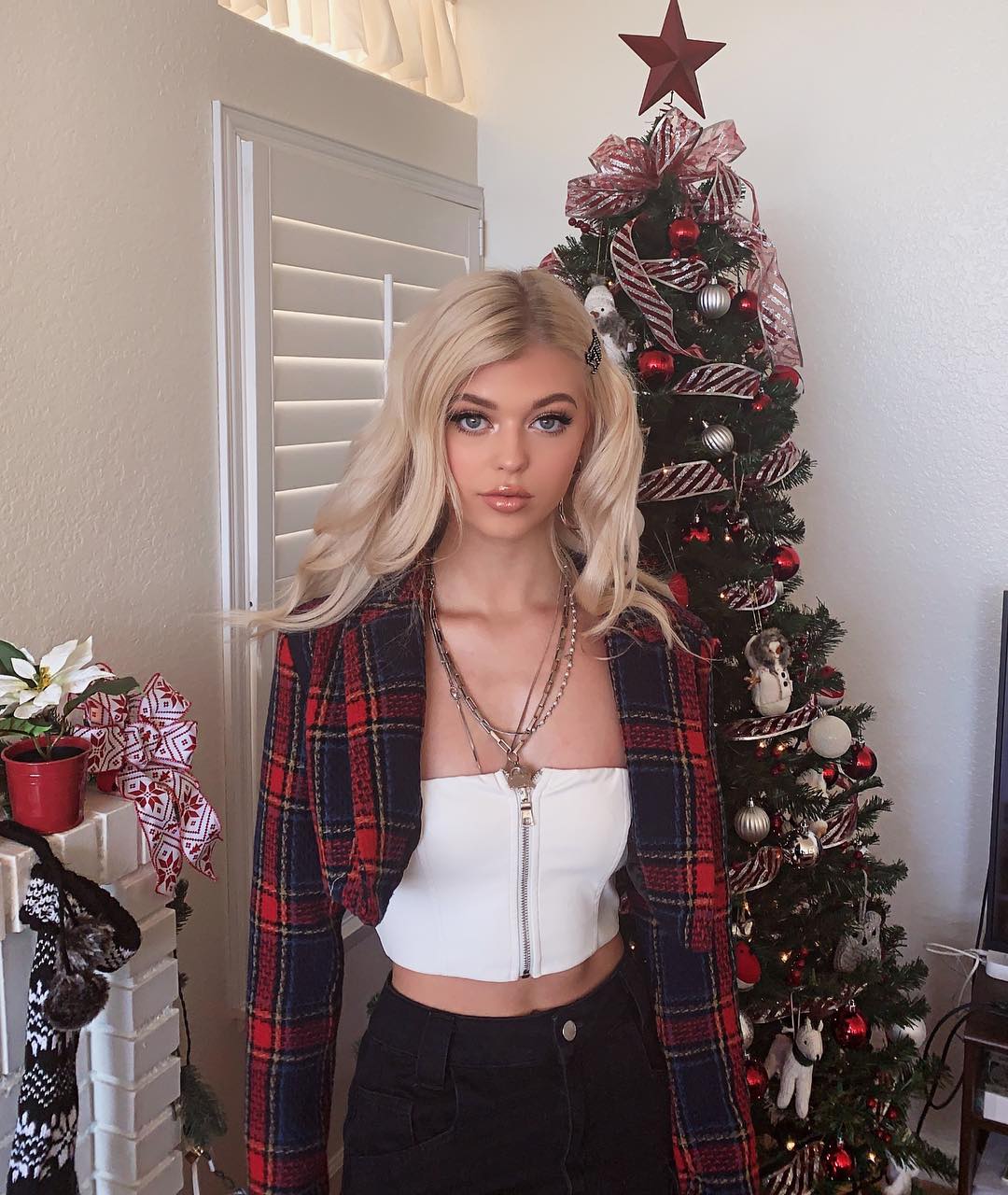 a woman with blonde hair is standing in front of a christmas tree. she is wearing a white crop top, black pants, and a plaid jacket. the christmas tree is decorated with a red star, ribbon, and ornaments. there is a mantle with a christmas stocking on it in the background.