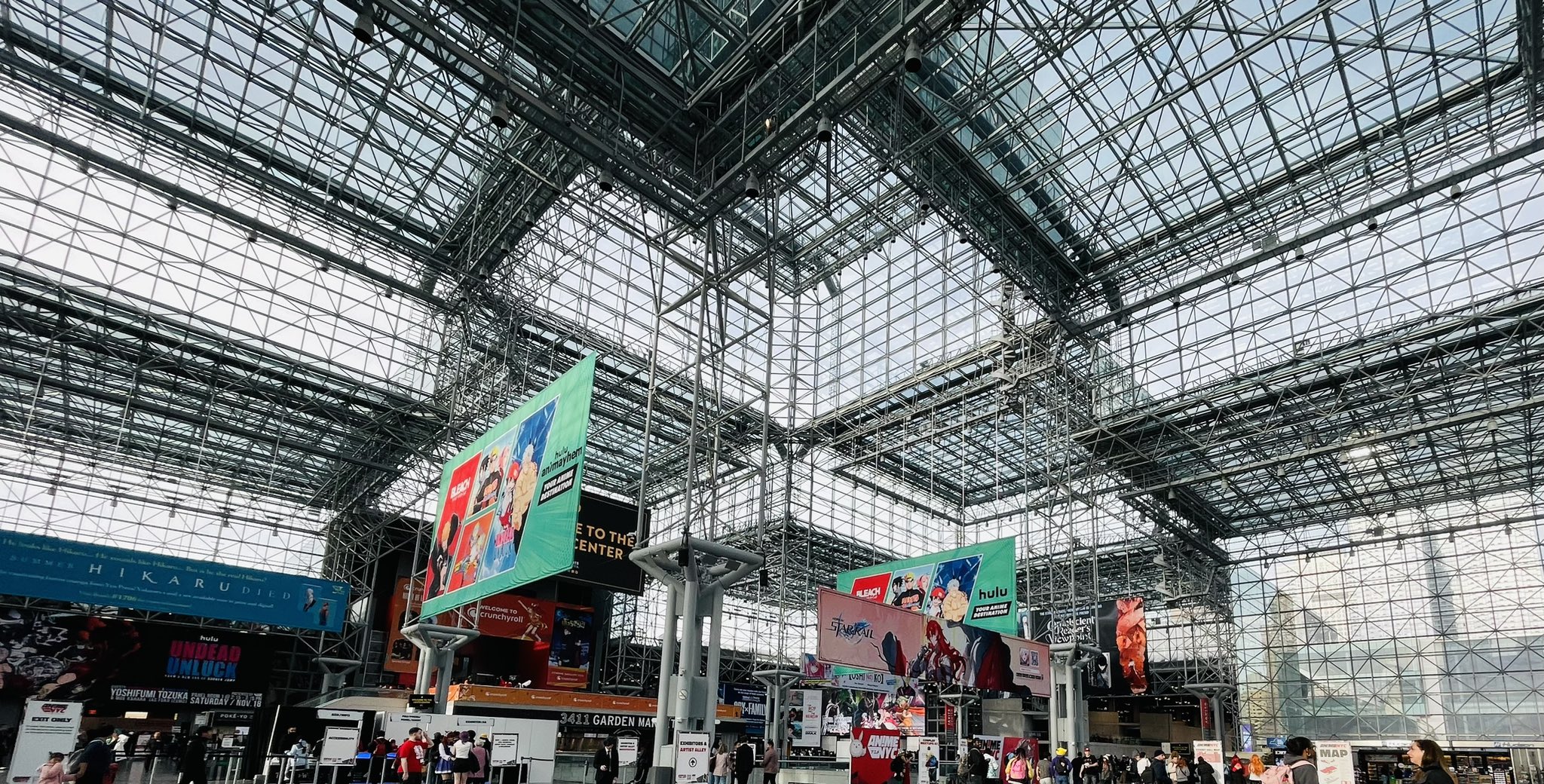 a wideangle shot looking up at the ceiling of a convention center. the ceiling is made of metal beams and glass panels, allowing natural light to flood the space.  large, colorful advertisements hang from the ceiling, showcasing various brands and products.  a crowd of people walk around below, adding to the bustling atmosphere of the event.  the overall impression is one of energy and excitement.