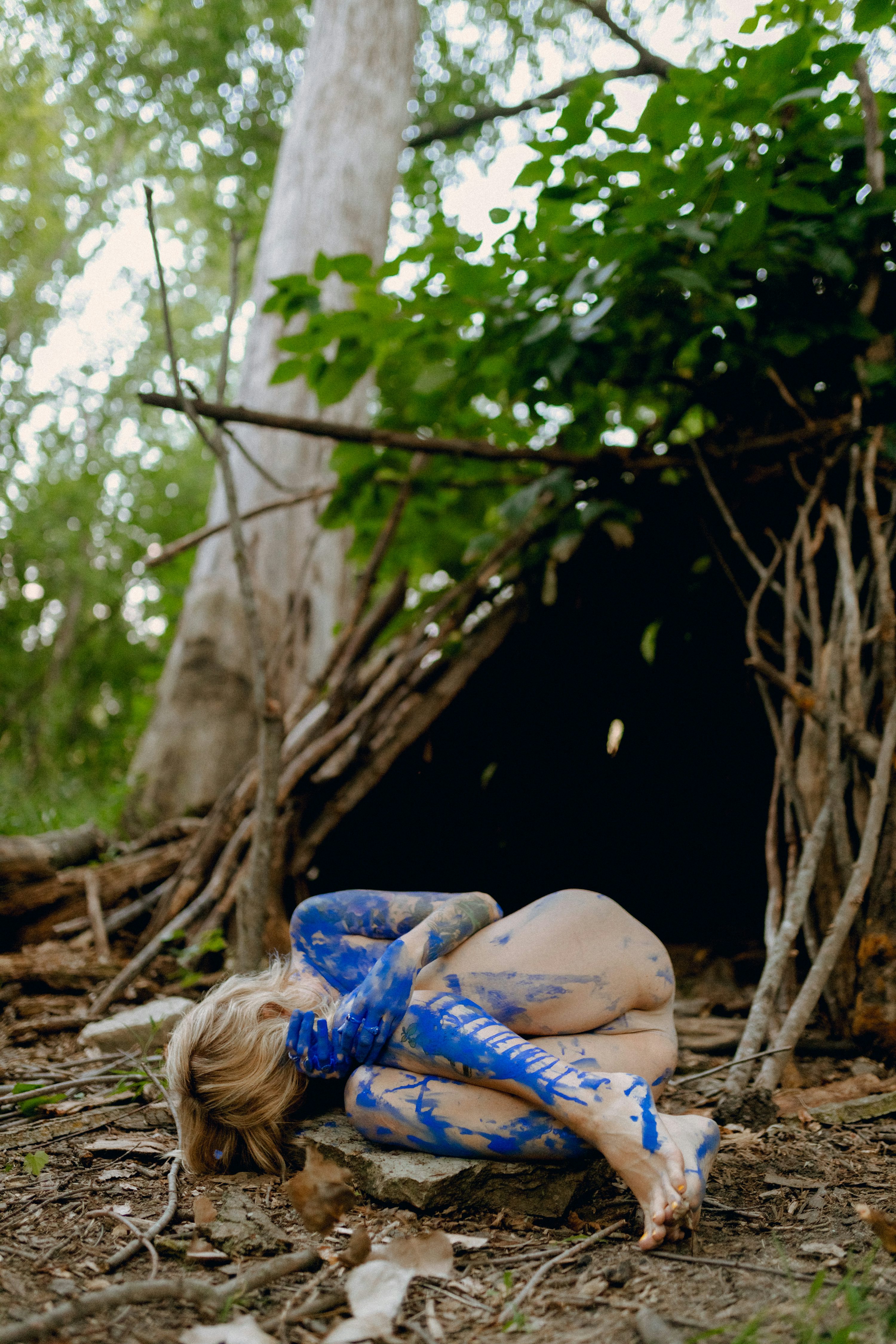 a woman with blue body paint is curled up on the ground in a secluded forest setting. she is near a simple leanto shelter made of branches and twigs. the overall mood is one of vulnerability and introspection. the image is artistic and evocative.