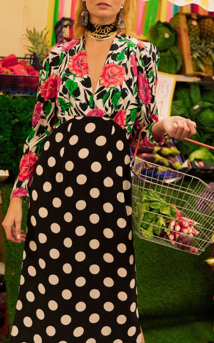 a woman in a stylish twotoned dress is shopping for groceries. the dress has a floral top and a black and white polka dot bottom. she is carrying a small shopping basket filled with vegetables and is wearing earrings and a necklace. the setting appears to be a vibrant and colorful outdoor market.