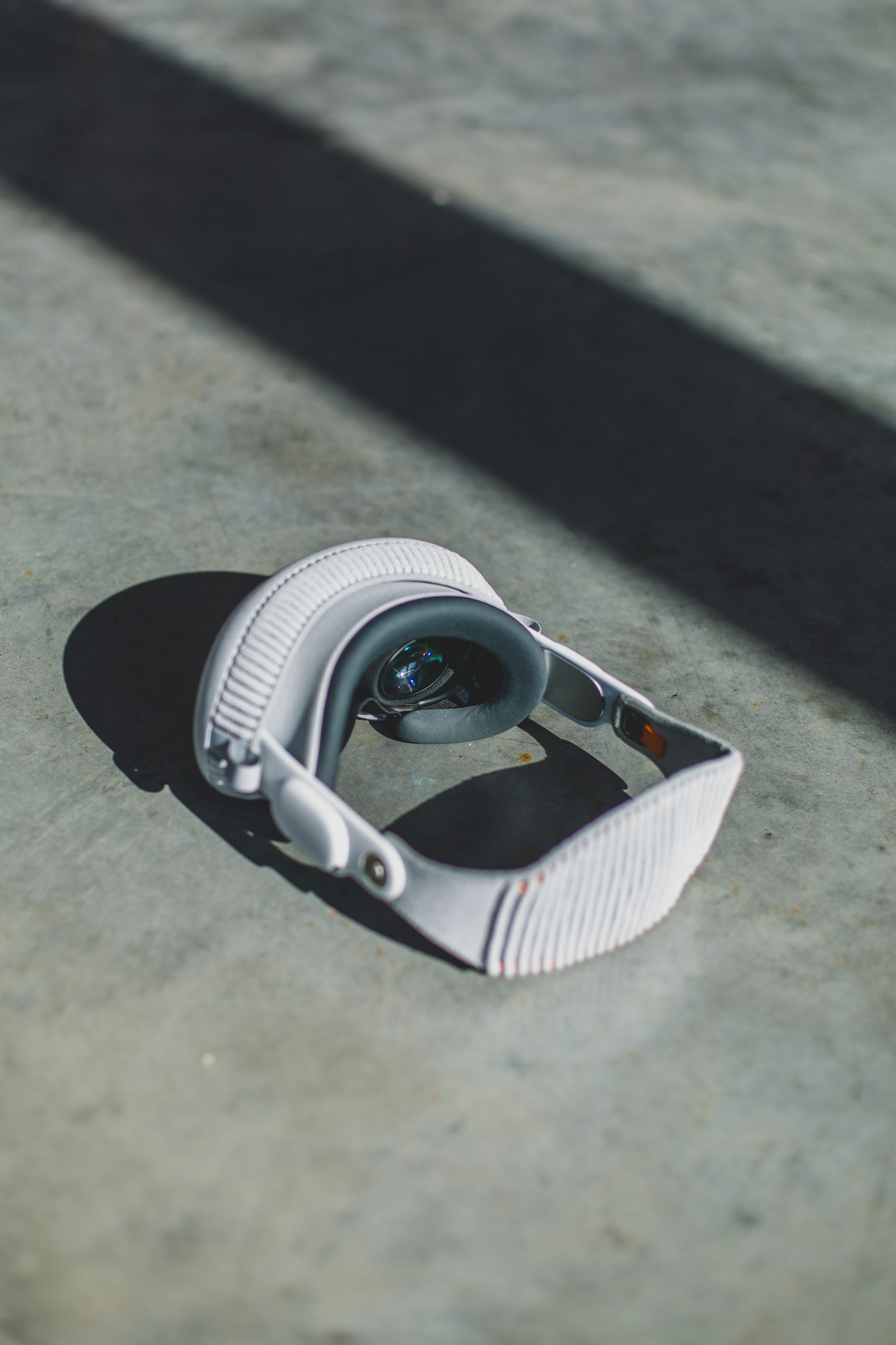 a white vr headset is lying on a grey concrete floor. there is a shadow behind the headset and a blurred shadow in the background. the image is indoors.