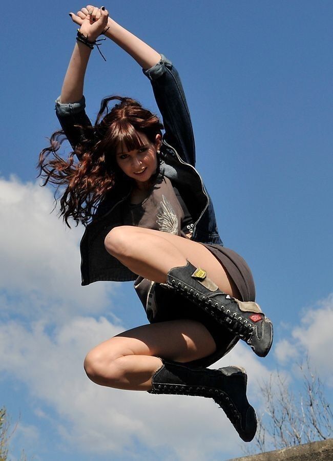 a woman in a jean jacket and boots is jumping in the air with her arms raised against a backdrop of a blue sky with clouds.