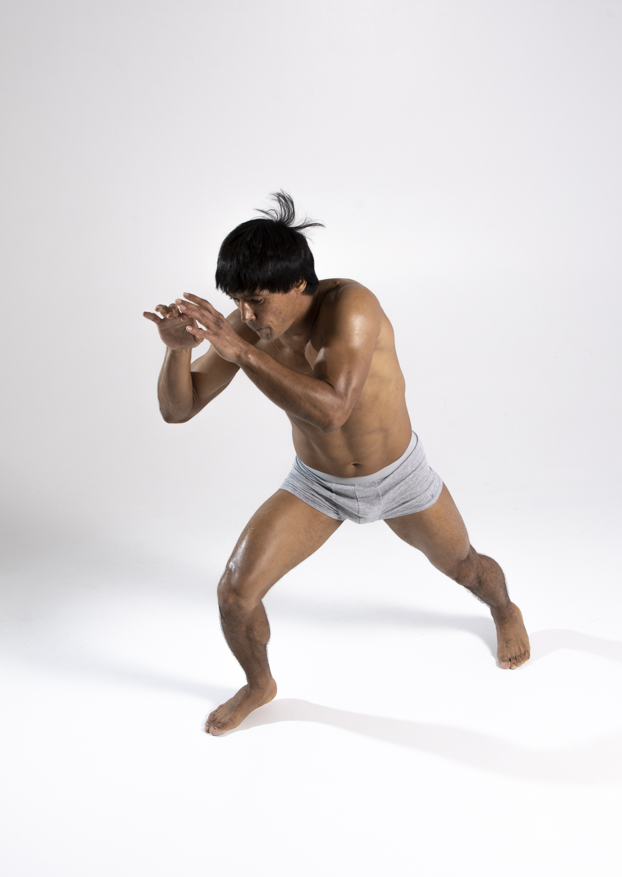 a muscular, shirtless man is depicted in a fighting stance against a plain white background. the man is wearing gray underwear and his skin appears to be oiled or sweaty, giving it a shiny sheen. he has dark hair that is styled with some strands sticking upwards. the man's body is toned and he is positioned with one leg forward and the other back, indicating movement or readiness for combat. his hands are raised in front of him, suggesting a defensive or offensive posture. the setting is a studio, evident from the seamless white backdrop.