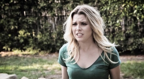 a young woman with blonde hair is standing outdoors in a garden setting. she is wearing a green vneck tshirt. she appears to be upset, frustrated, or angry, as evidenced by her furrowed brow and tense facial expression. the lighting is natural and slightly overcast, suggesting it might be a cloudy day. the perspective of the shot is at eye level.