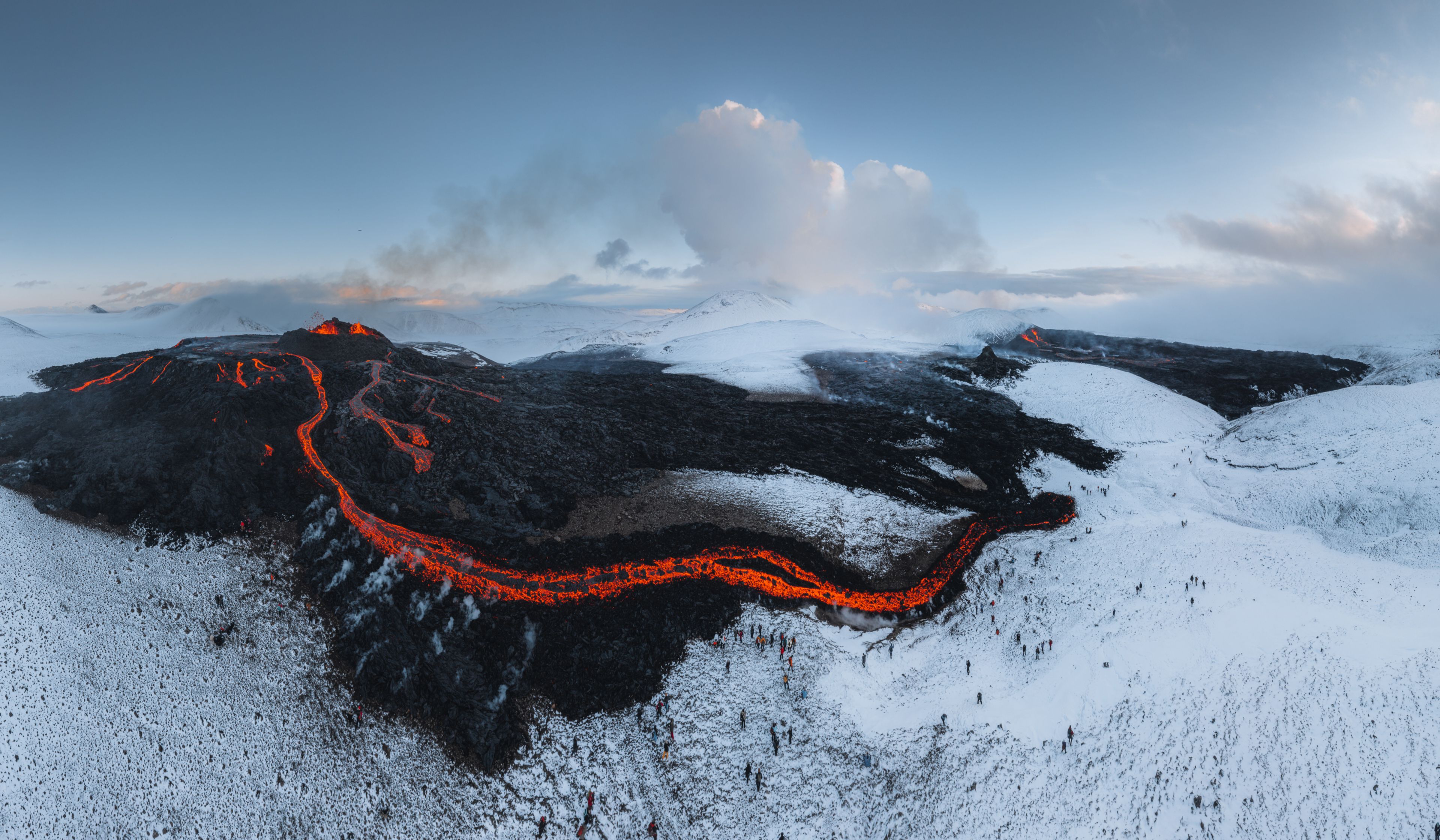 an aerial view of a volcano erupting in iceland. the lava flows down the side of the mountain and across the snowcovered landscape. the sky is blue and clear, and the sun is shining. the contrast between the hot lava and the cold snow is stark and dramatic.