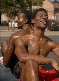 the image shows two muscular black men sitting on a basketball court on a sunny day. they are both shirtless and covered in glitter, with big smiles on their faces. the man in the foreground is wearing red athletic shorts.  the image evokes feelings of joy, friendship, and summer fun.