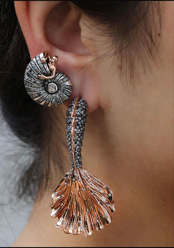 a closeup shot of a woman's ear adorned with ornate earrings. the first earring is a nautilus shell design, featuring a small figure of a person seemingly embracing the shell. it has a combination of silver and rose gold, with diamonds embedded throughout. the second earring is a mermaid tail design with a rose gold and silver color scheme with diamonds throughout.