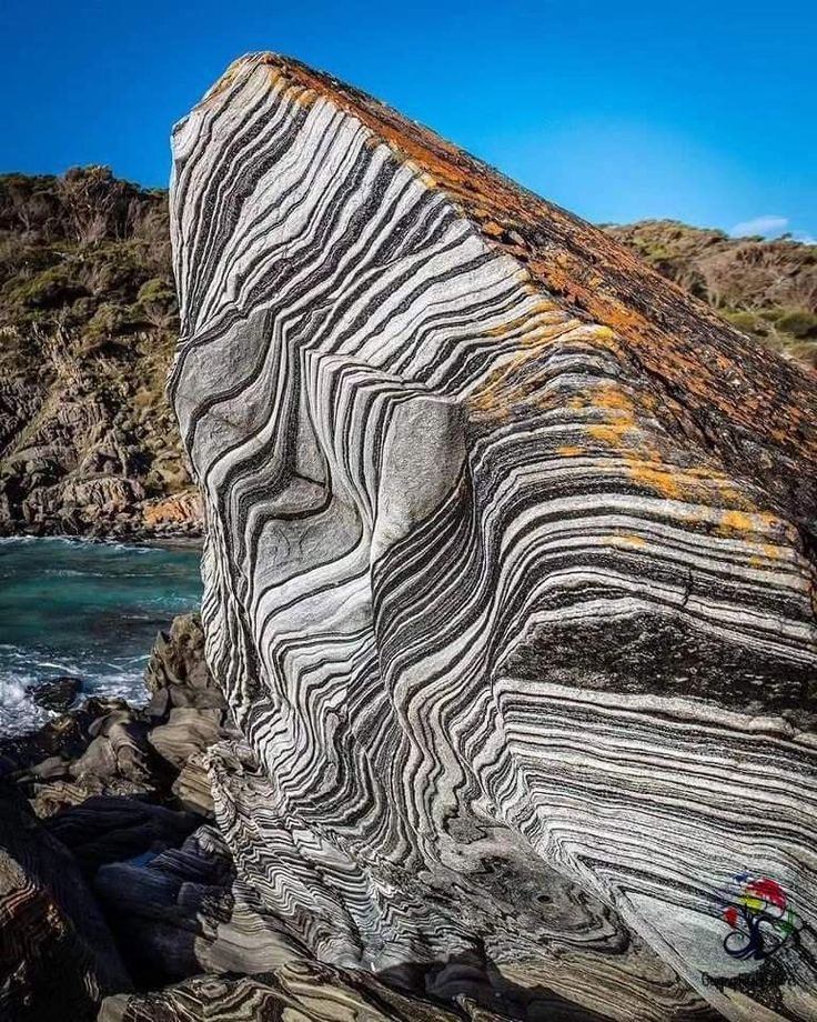 a large striped rock formation stands tall against a backdrop of the ocean and a rocky shore. the rock's surface is characterized by a mesmerizing pattern of black and white stripes, created over millennia by geological processes. the ocean's waves crash against the shore, highlighting the rock's dramatic presence and the relentless forces of nature. the overall scene is a testament to the power and beauty of the natural world.