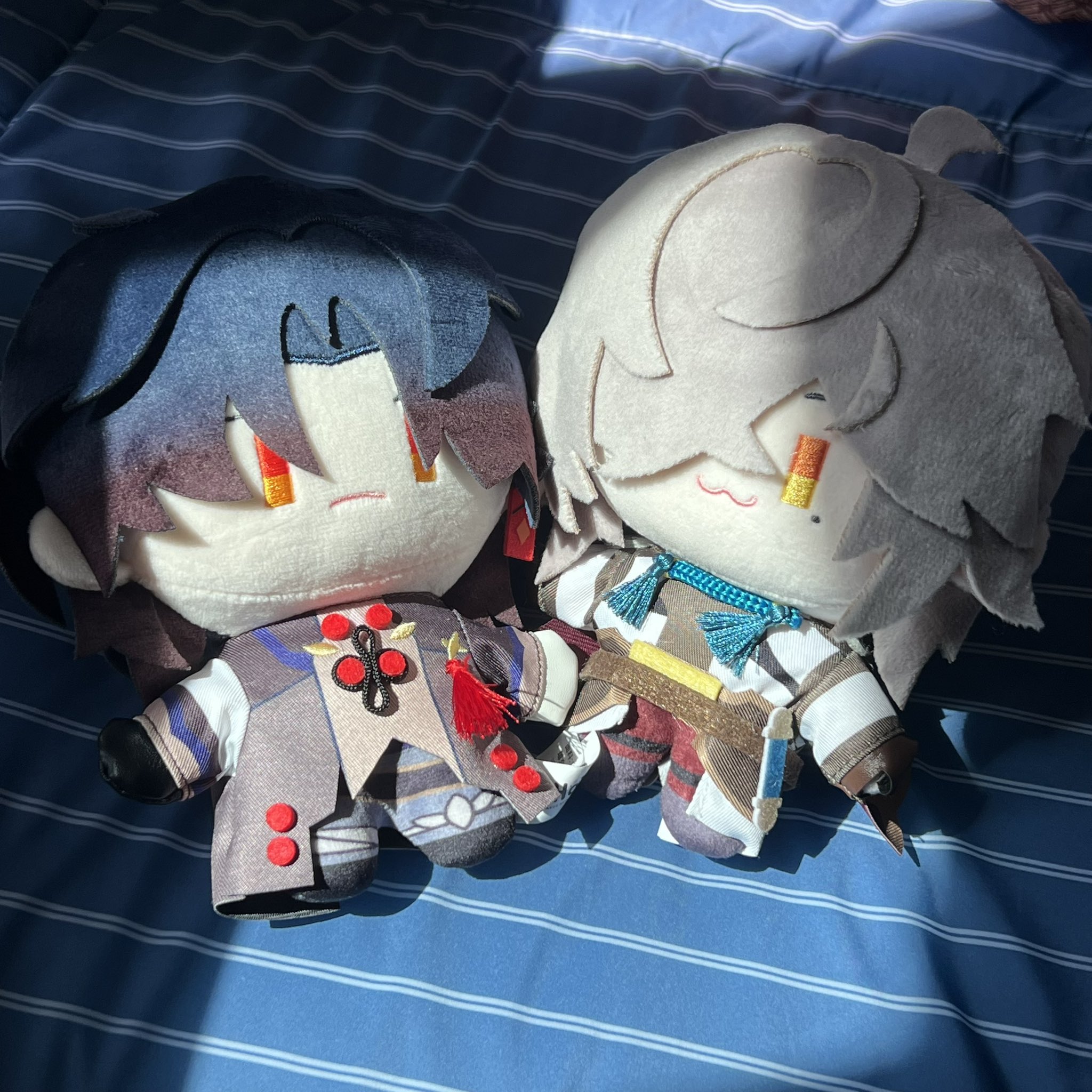 two plush anime characters sit on a blue and white striped background. one is a character with black hair, red eyes, and a black and red outfit. the other is a white haired character with red eyes, and a white and brown outfit.
