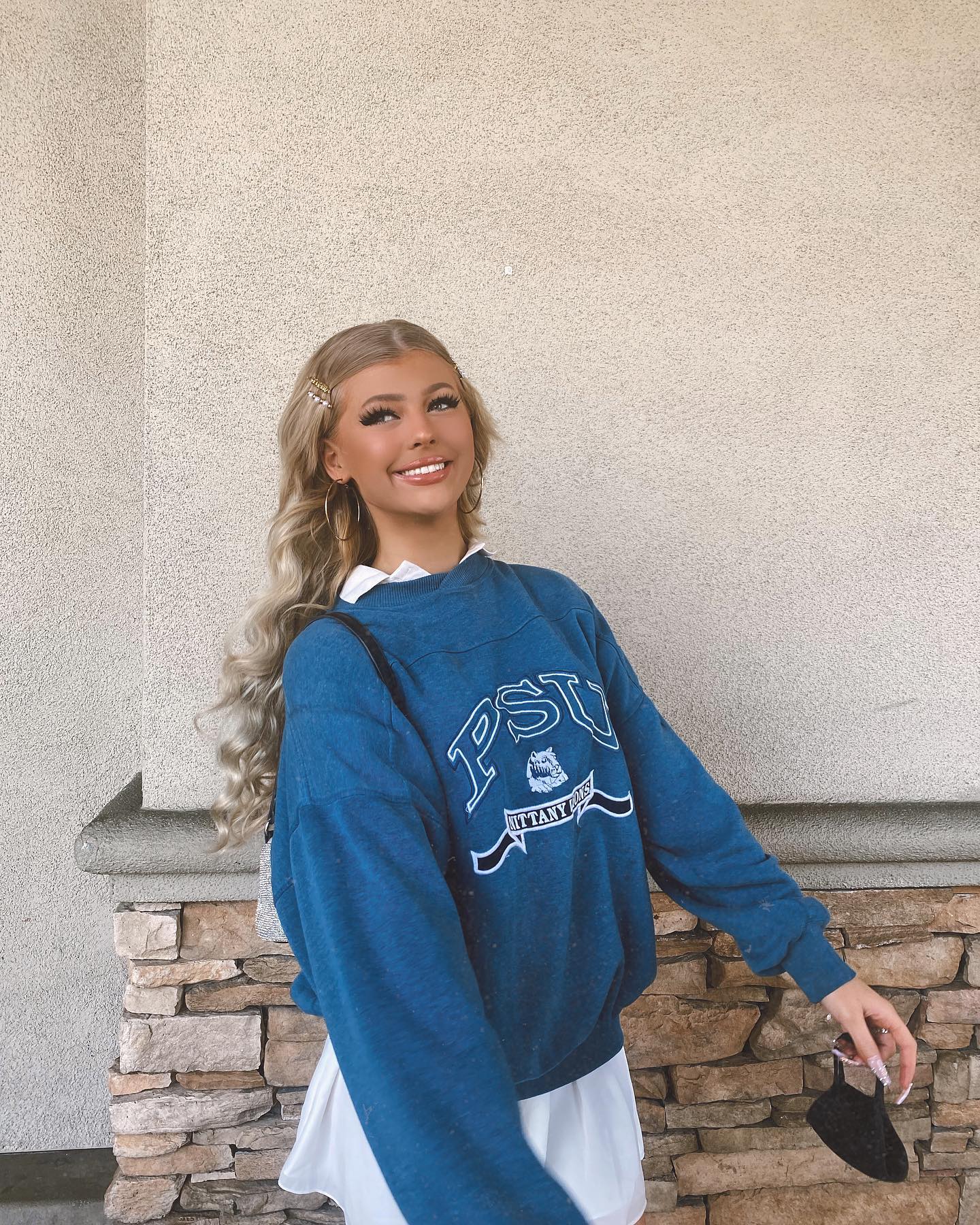 a woman with blonde wavy hair is smiling and wearing a blue penn state university sweatshirt over a white dress. she has hair clips in her hair and is holding a black face mask in her right hand. she is standing in front of a brick wall.