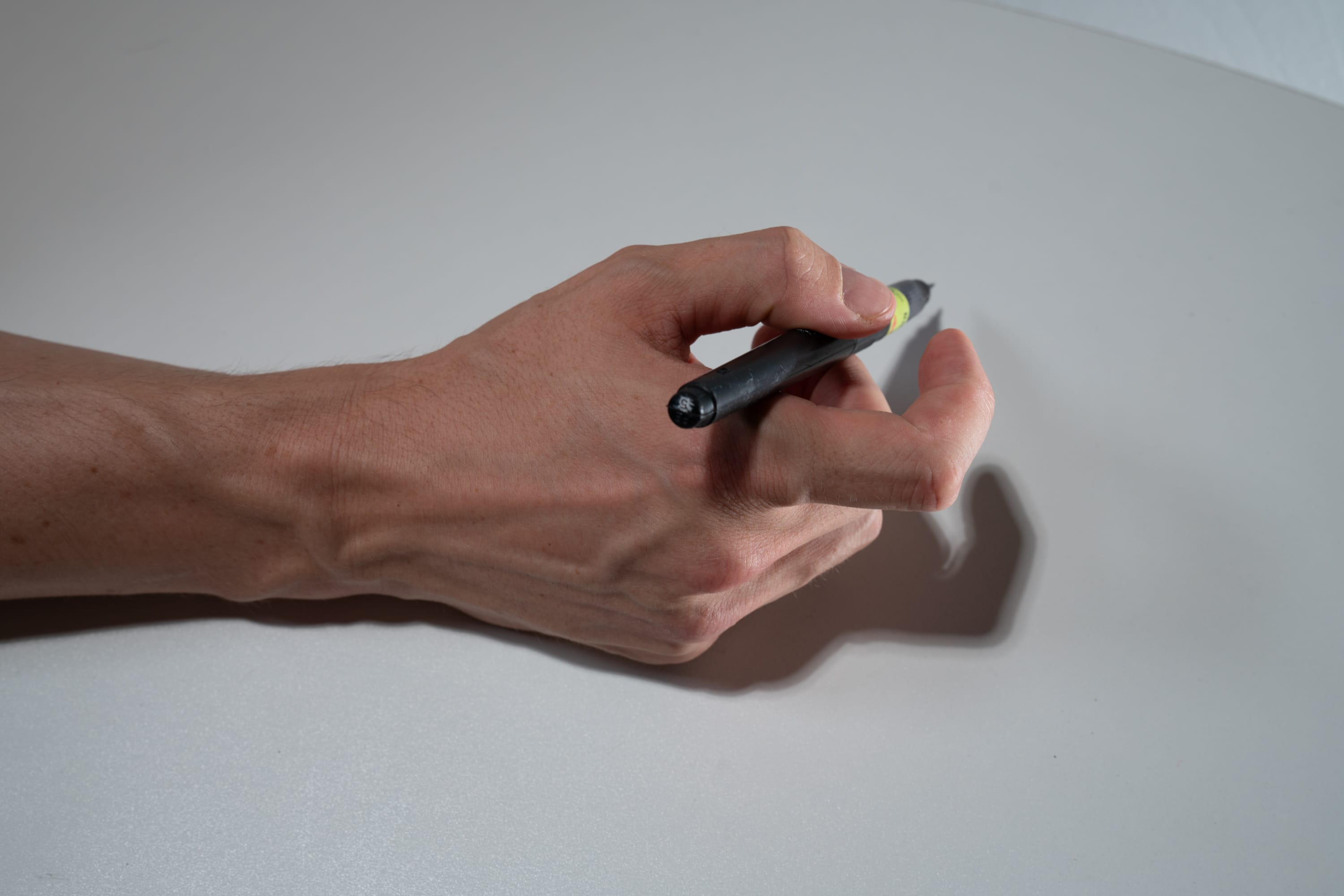 a man's hand holding a black pen is captured from a topdown perspective against a white background. the image highlights the details of the hand's skin and prominent veins, while a subtle shadow cast by the hand adds depth to the composition.