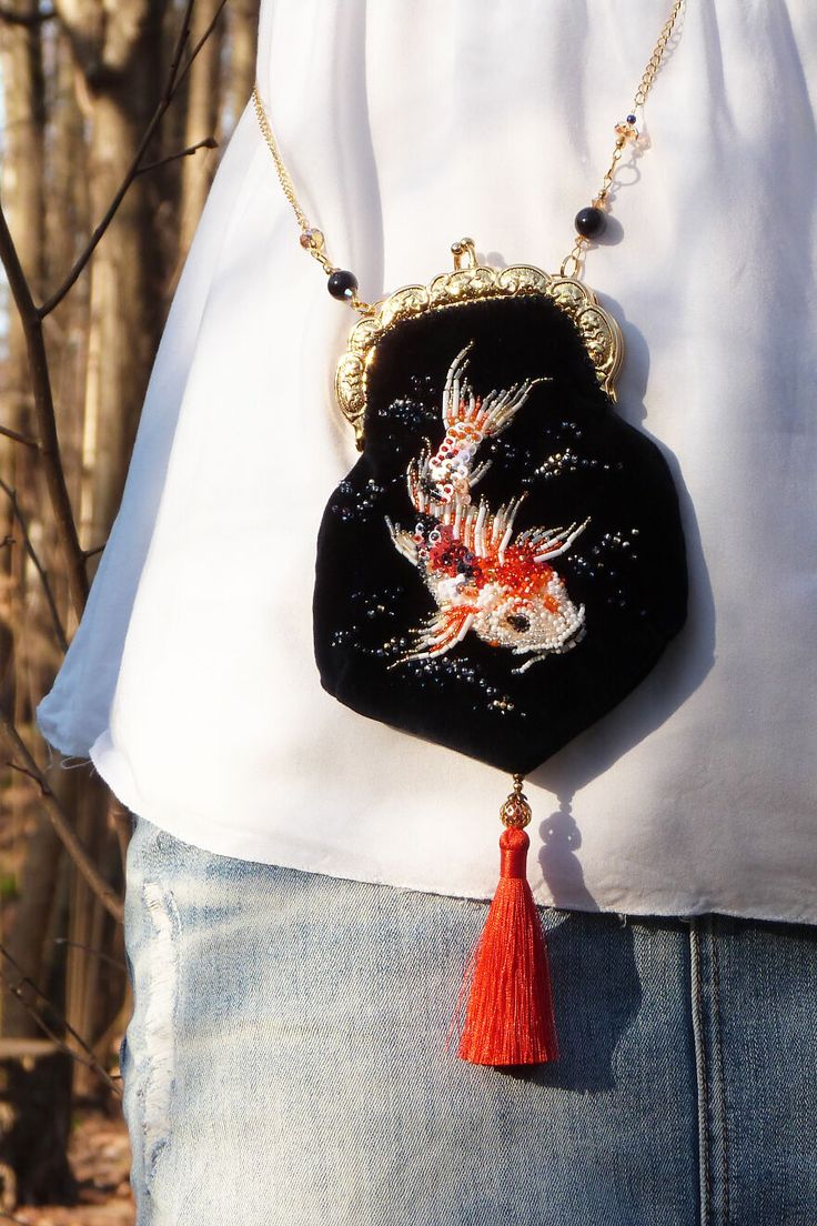 a woman is wearing a white shirt with a black bag and a gold chain. the bag has a red tassel. there is an embroidered image of a beaded fish on the bag. the woman is wearing blue jeans. the woman is standing in front of a tree.