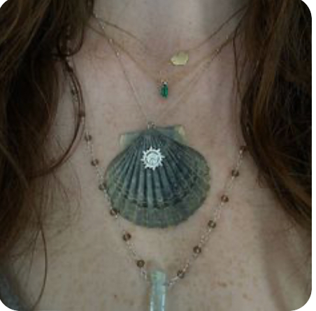 a closeup photo showcasing multiple delicate necklaces on a woman. the focal point is a large scallop shell adorned with a sun pendant. other necklaces feature a green stone and a small gold charm. the overall aesthetic exudes a boho, beachy vibe.