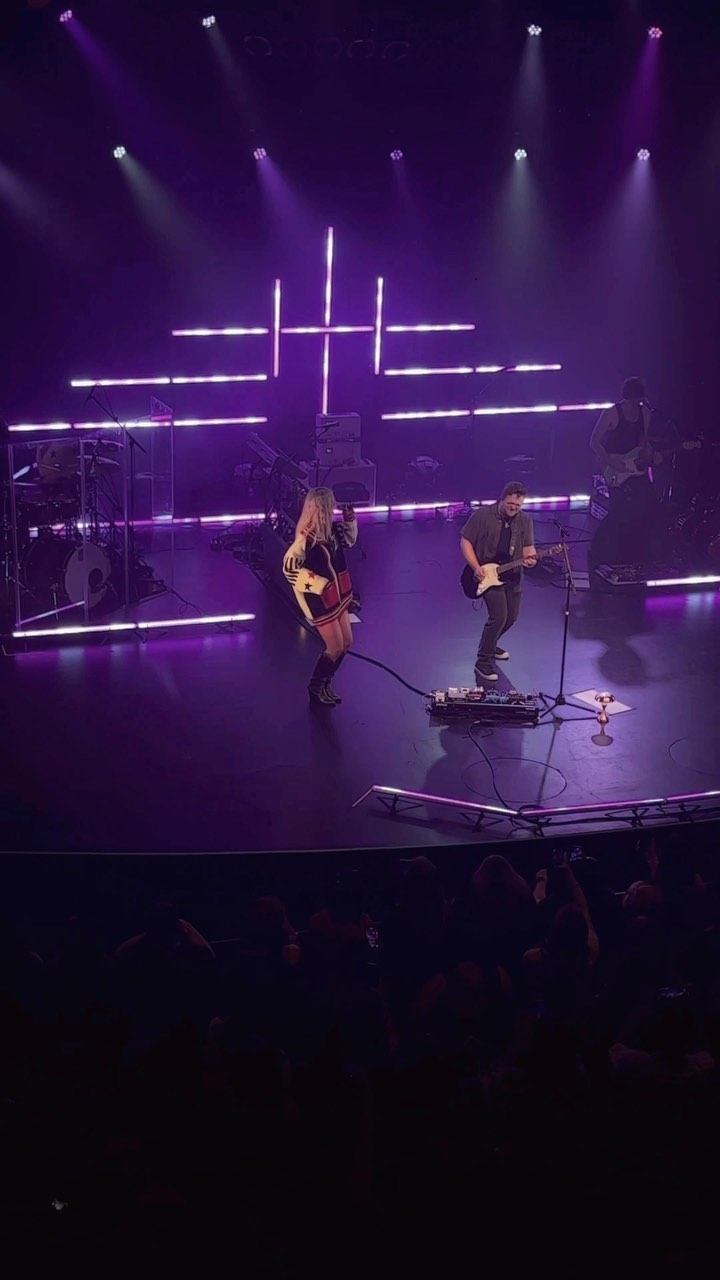 a concert stage is lit up with spotlights, featuring musicians performing for a large audience. a woman in a short skirt and long boots is singing with a guitar, accompanied by a man playing an electric guitar. the stage has a backdrop of illuminated lines, and the audience is visible in the lower part of the frame.