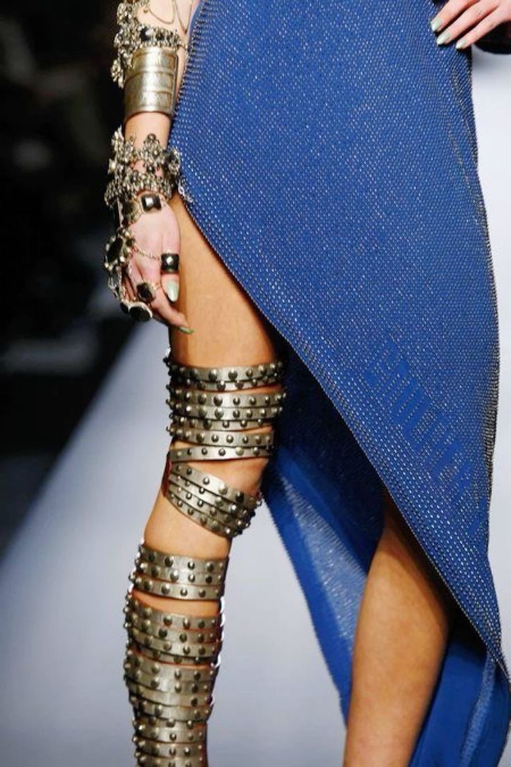 a fashion model walks the runway wearing a blue dress with a thigh high slit and silver studded boots. she has silver bracelets on her arms and rings on her fingers. the model's hair is styled in a sleek updo, and she is wearing minimal makeup. the focus of the photo is on the model's legs and the boots. the photo is taken from a low angle, and the background is blurred, making the model the focal point of the image. the photo is a good representation of the latest trends in fashion.