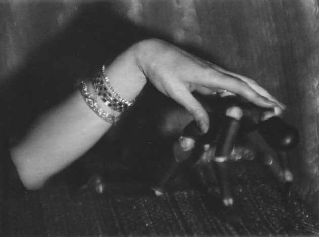 a black and white closeup shot of a woman's arm adorned with multiple bracelets. her hand rests on a dark, seemingly antique object, possibly a piece of machinery or art, that's partially obscured by shadows. the image evokes a sense of mystery and intrigue, with the focus on the contrast between the woman's delicate jewelry and the rough texture of the object she's touching. the overall aesthetic is vintage and artistic, with a strong emphasis on shadow and light.