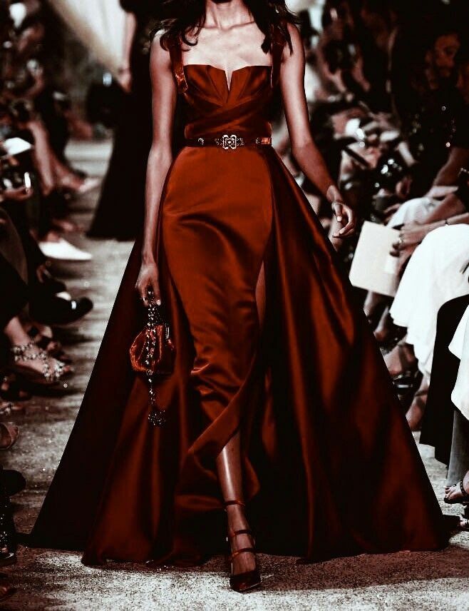 model walks down a runway in a stunning deep red, almost burgundy, satin gown. the dress features a fitted bodice with a sweetheart neckline, a highwaisted belt with ornate detailing, and a dramatic long train that flows behind her.  the model's legs are partially visible through a high slit in the skirt. she carries a small, darkcolored handbag and wears elegant heels. the background is blurred but shows a packed audience watching the fashion show. the overall aesthetic is dark, moody, and luxurious. the image has a highfashion, editorial feel.