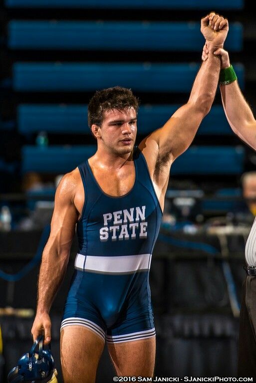 a penn state wrestler raises his arms in victory after winning a match. the wrestler is wearing a blue singlet with a white stripe and the words "penn state". he is smiling and appears to be very happy with his victory.  the picture was taken in a wrestling arena, and the background is blurred.