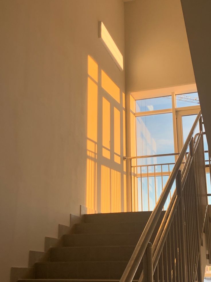 a close up of an interior staircase, showing the wooden steps, metal handrail and  the shadows of the large windows that cast a warm light. the background shows the rest of the interior, which appears clean and minimal. the stairs are welllit, giving the scene a bright and inviting feel.  the image is captured from a low perspective, creating a sense of depth and upward movement.