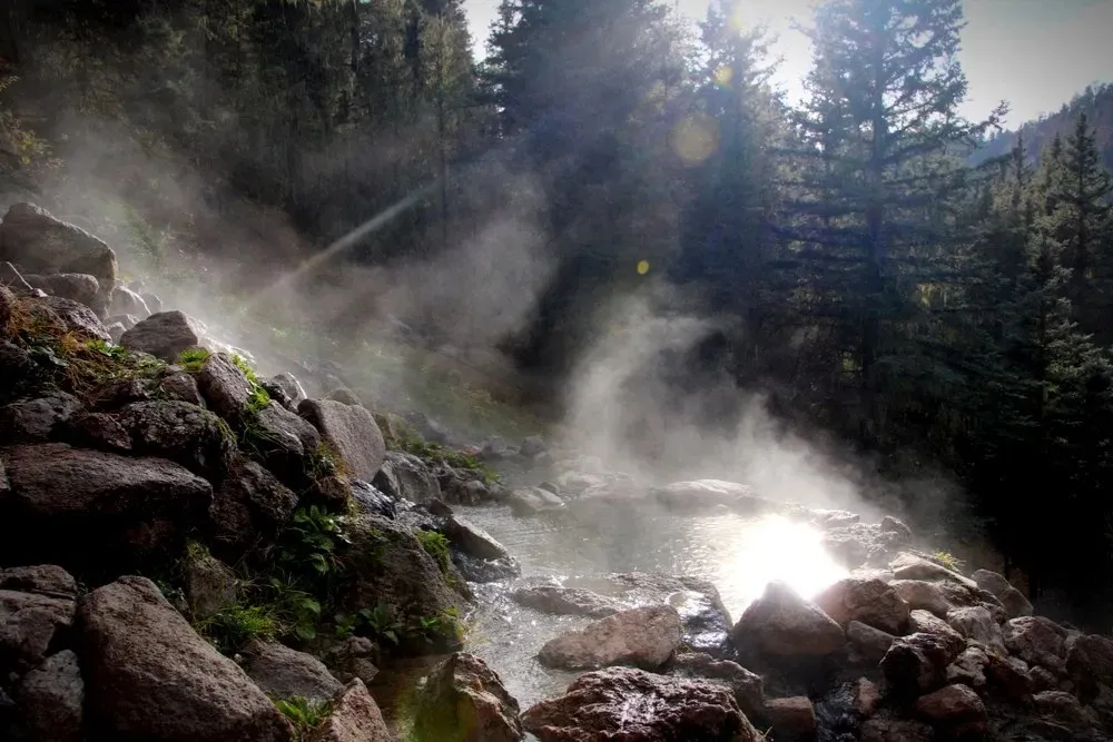the image shows a natural hot spring surrounded by rocks and trees. steam rises from the water, illuminated by sunlight filtering through the forest. moss and vegetation grow on the rocks, adding to the natural beauty of the scene.