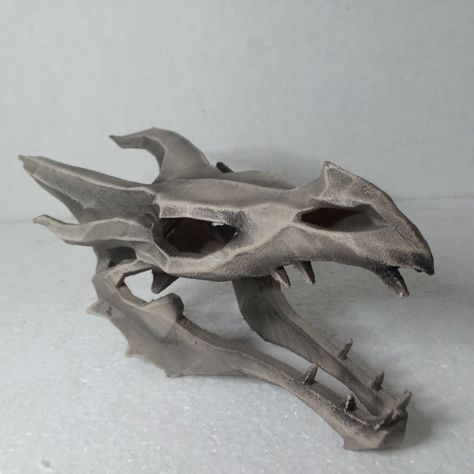 a closeup image of a 3d printed model of a dragon skull.  the skull is painted in a light gray color, with a rough texture that gives it a realistic look. the skull is positioned on a white surface.  the image is welllit and the dragon skull is the only object in the image.