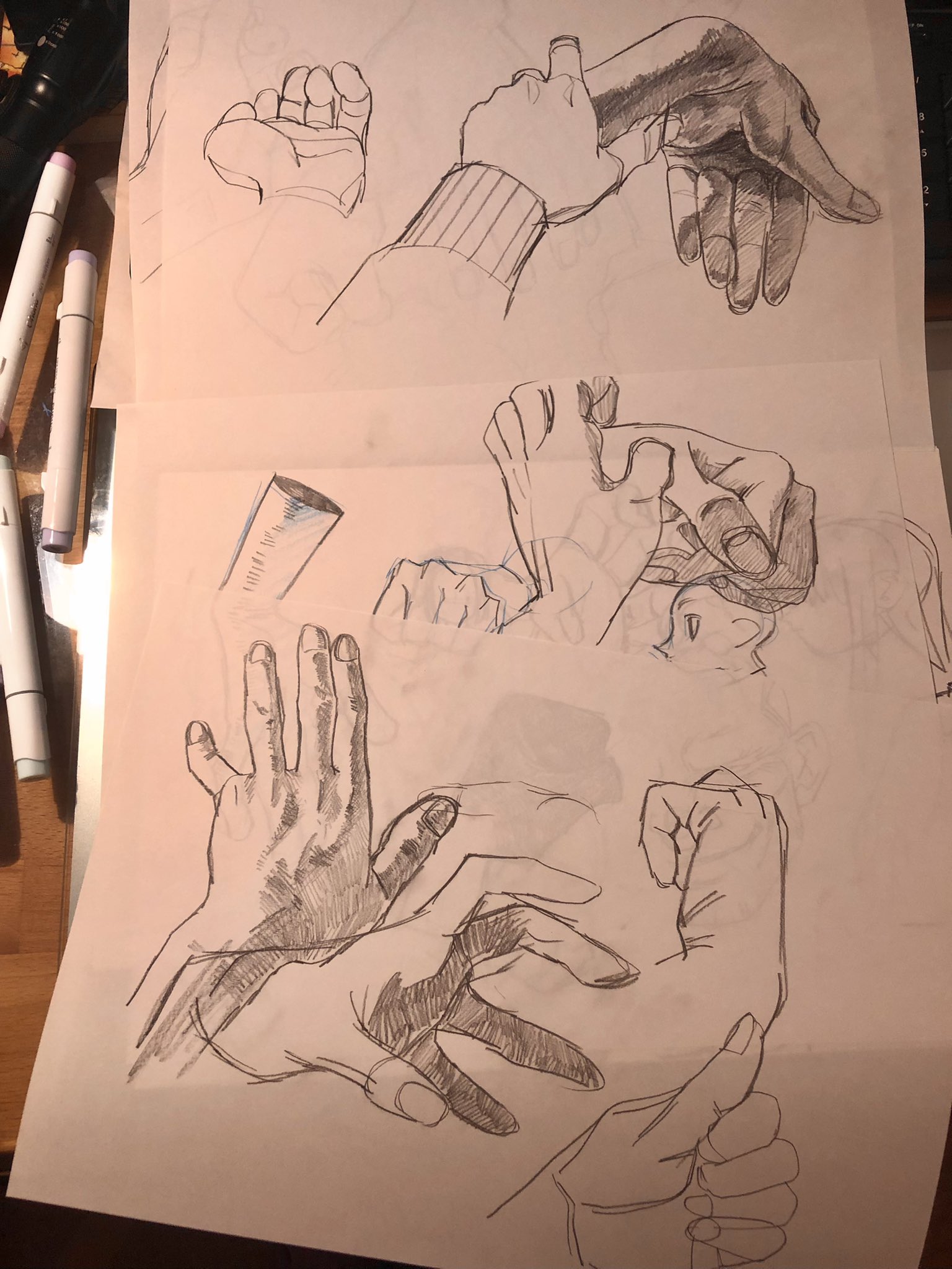 the image shows several pieces of paper with pencil sketches of hands in different positions and perspectives. the sketches appear to be part of an artist's anatomy study, focusing on understanding the structure and movement of hands. the drawings range in levels of completion, with some being more detailed with shading while others are primarily outlines. the use of light and shadow suggests the artist is paying attention to form and volume.  on the left side of the image, there is a set of art markers, indicating the artist's tools.