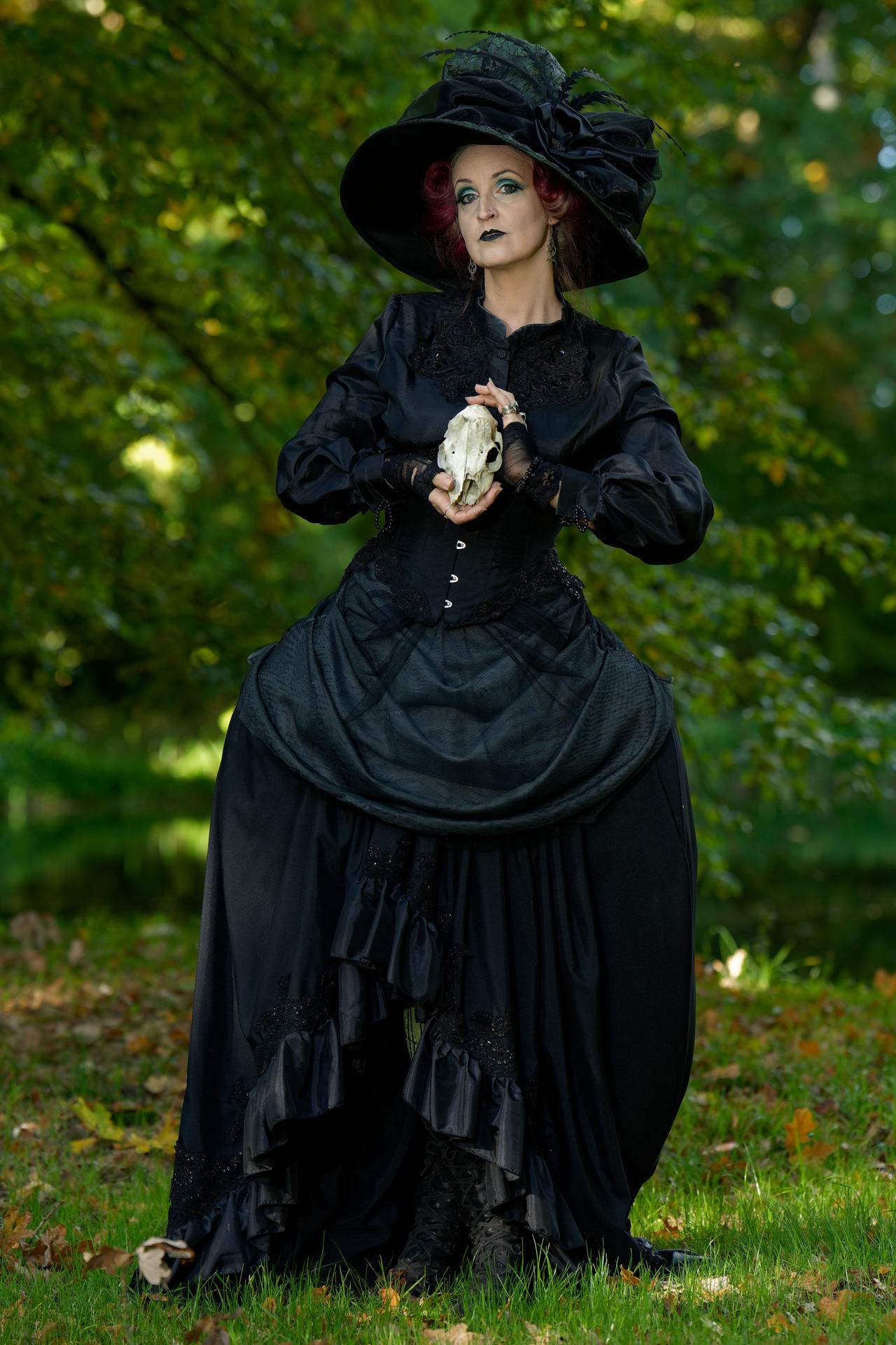 a woman dressed as a gothic witch stands in a forest clearing. she wears a long, black, ruffled dress with a corset, a large black hat with feathers, and black gloves. her makeup is dark and dramatic. she holds a small skull in her hands. the background is a blur of green trees and grass.