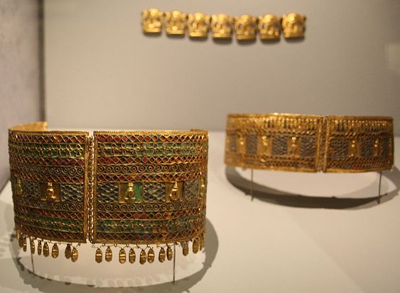two ancient greek gold necklaces are displayed in a museum.  the necklaces are decorated with intricate patterns and colorful gems. they are mounted on stands for display.