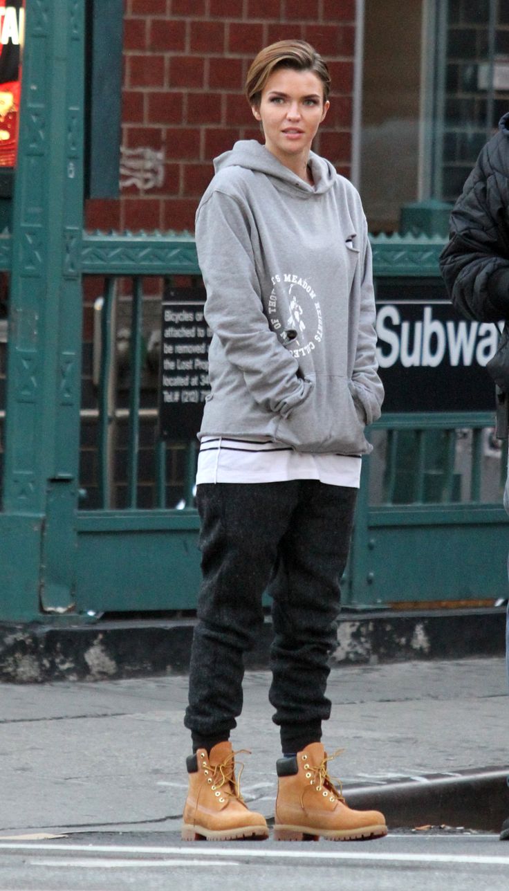 ruby rose is pictured on a new york city street, wearing a grey hoodie, black sweatpants, and timberland boots.  she's casually dressed, her short hair styled simply.  a portion of a subway entrance is visible in the background. the overall setting suggests a cool and casual autumn or winter day.