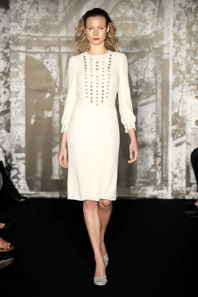 a full shot of a fashion model walking down a runway. she is wearing a simple, elegant white midi dress with gold studding along the front. the dress is longsleeved and has a straight silhouette. the model has blonde curly hair and is wearing simple grey flats. the background is a blurred image of a grayscale wall.