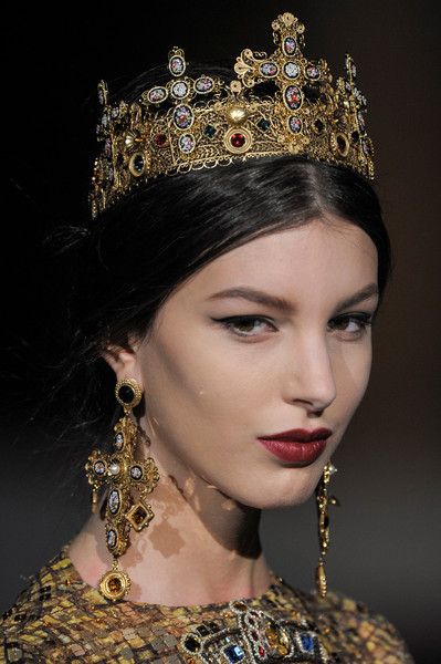 loseup view of a fashion model wearing a magnificent gold crown adorned with intricate details, including crosses and colorful gemstones.  she also sports matching large crossshaped earrings with similar ornate detailing.  her makeup is dramatic with dark eyeliner and a deep burgundy lipstick.  she is wearing a richly textured gold and brown patterned outfit. the overall style is regal and opulent, suggesting a highfashion runway show, possibly by dolce & gabbana. the background is dark and blurred, focusing attention on the model and her jewelry.