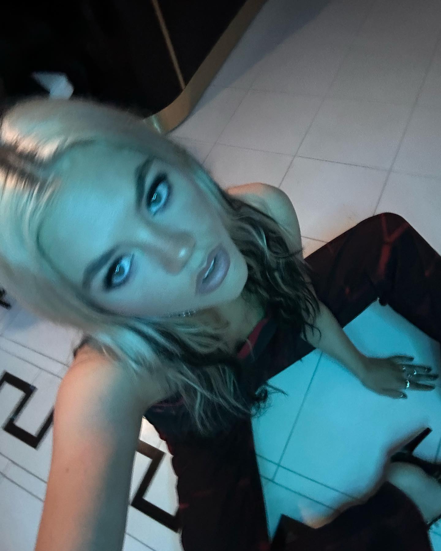 a woman with blond hair and blue eyes is sitting on a black and white tiled floor. she is wearing a dress and rings. the image is lit with a blue light.