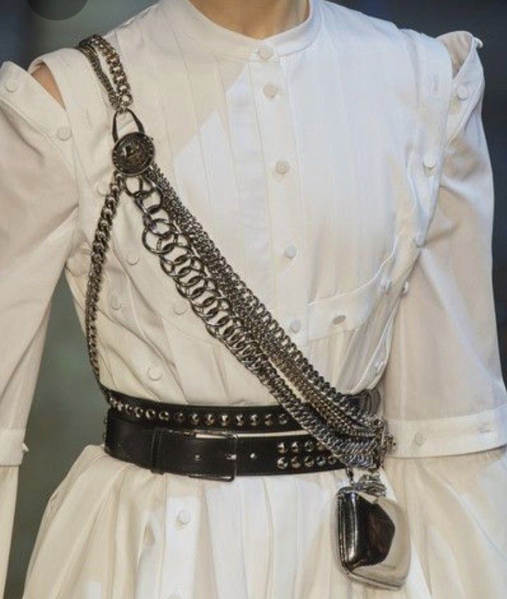 a white dress is shown with a black belt around the waist and multiple silver chains draped over the shoulder and across the body. the dress has buttons down the front, an open shoulder on one side, and a pocket. the black belt has a silver buckle and studs.