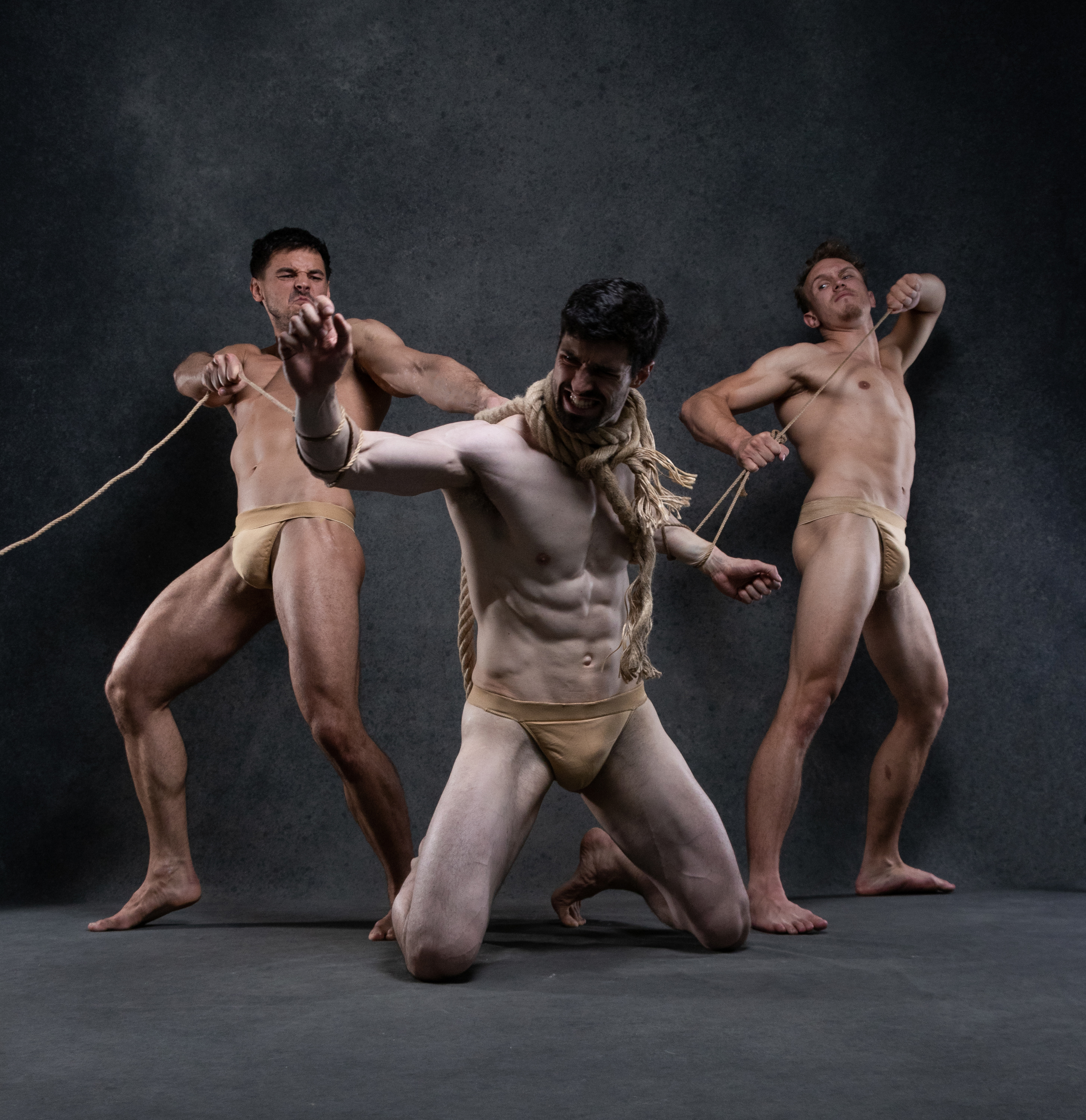 three muscular men, wearing only beige loincloths, are engaged in a tense struggle with a thick rope against a dark gray backdrop. the man in the center, illuminated by dramatic lighting, appears to be the focal point of the composition, his face contorted in effort. the other two men flank him, their bodies taut with tension as they pull on the rope.