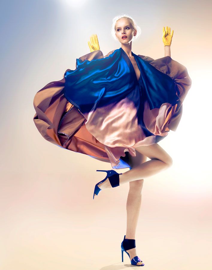 this image is a fashion photograph of a model. the model is a young woman with long blonde hair and a light complexion. she is wearing a blue, pink, and purple dress with long, flowing sleeves. the dress is made of silky material. she is also wearing yellow gloves and black shoes. the background is light with white and pink tones.  the model is posing and looking at the camera, with her arms outstretched. she is standing on one leg and balancing on her other leg, with her leg bent and foot pointing to the side.