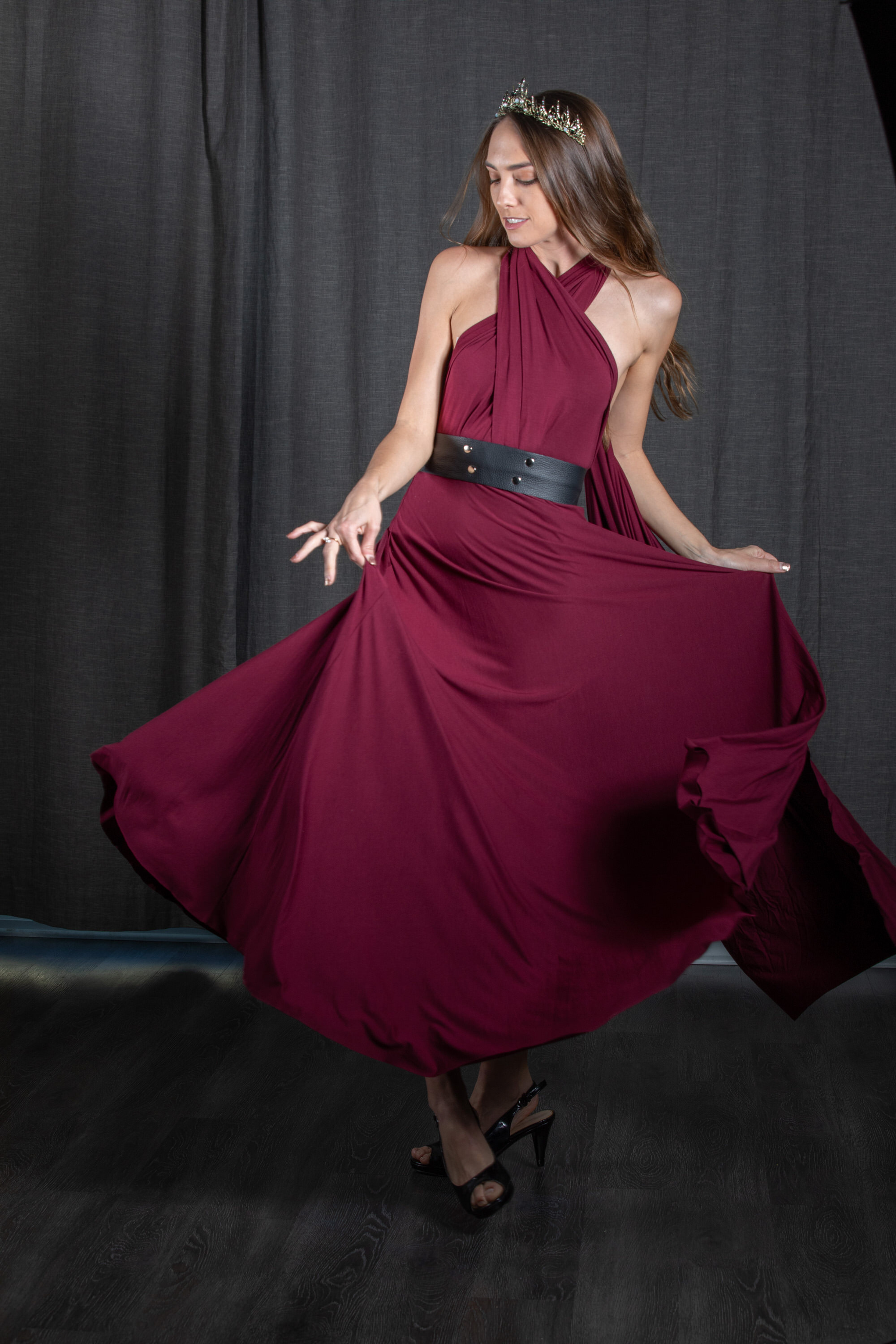 a young woman in a flowing burgundy dress dances gracefully against a dark backdrop. she wears a delicate crown, and her pose highlights the movement of the fabric. the studio setting and soft lighting contribute to an air of elegance and sophistication.