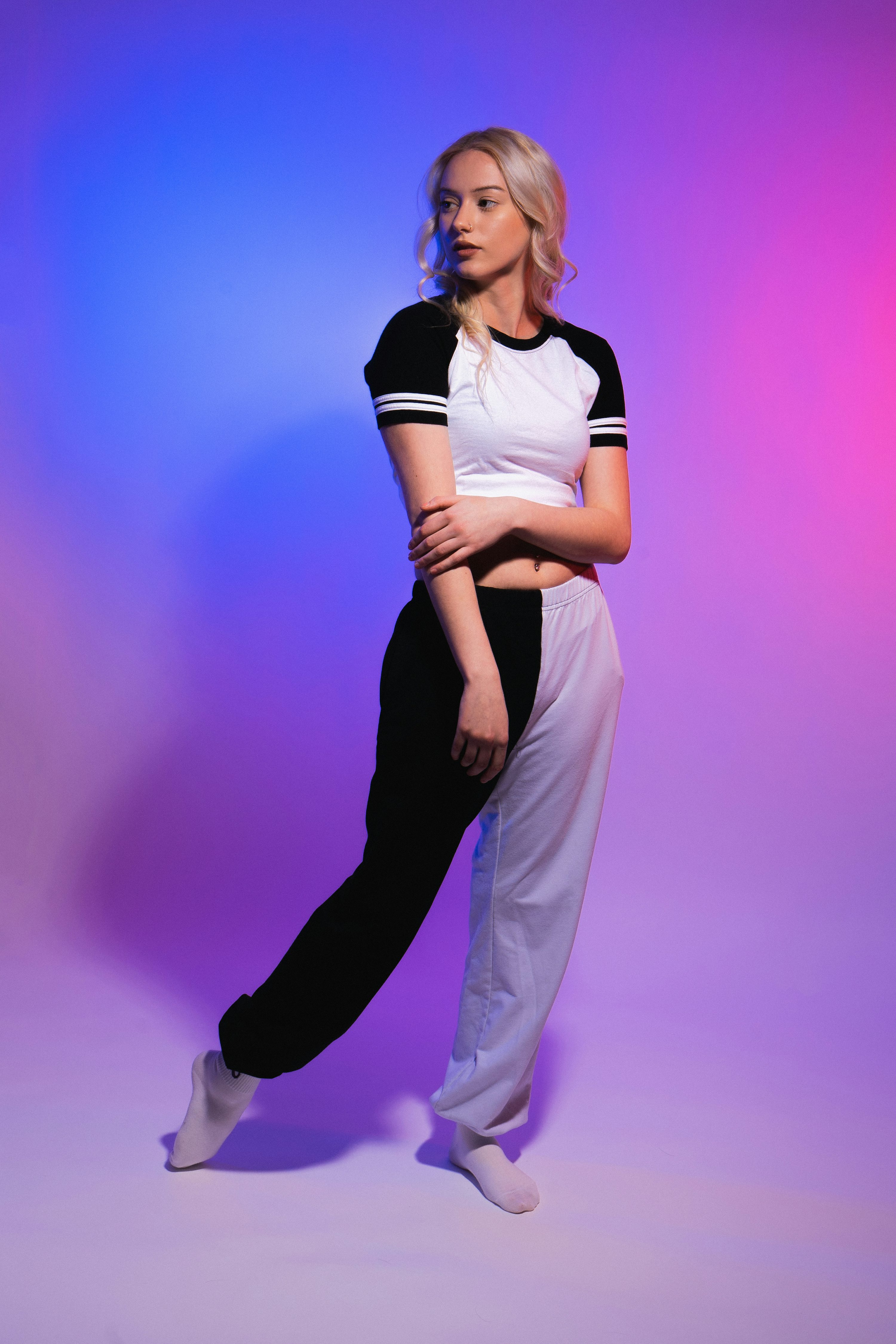 a fulllength shot of a young blonde woman model posing in a studio with a purple and blue gradient background. she's wearing a black and white twotoned pair of trousers, a white and black baseballstyle crop top, and white socks. her pose is relaxed yet stylish, and she looks away from the camera. the image has a trendy, contemporary feel with vibrant lighting.